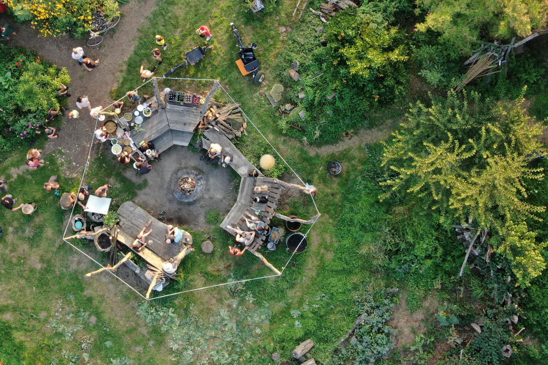 Luftaufnahme eines üppigen Gartens mit angrenzenden Gebäuden, in dem sich Menschen auf Bänken um ein Lagerfeuer versammeln