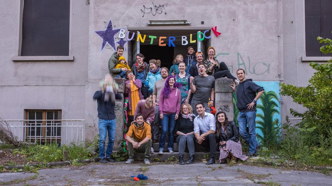 Eine bunt gemischte Personengruppe hat sich vor einem Hauseingang versammelt und blickt jubelnd zur Kamera